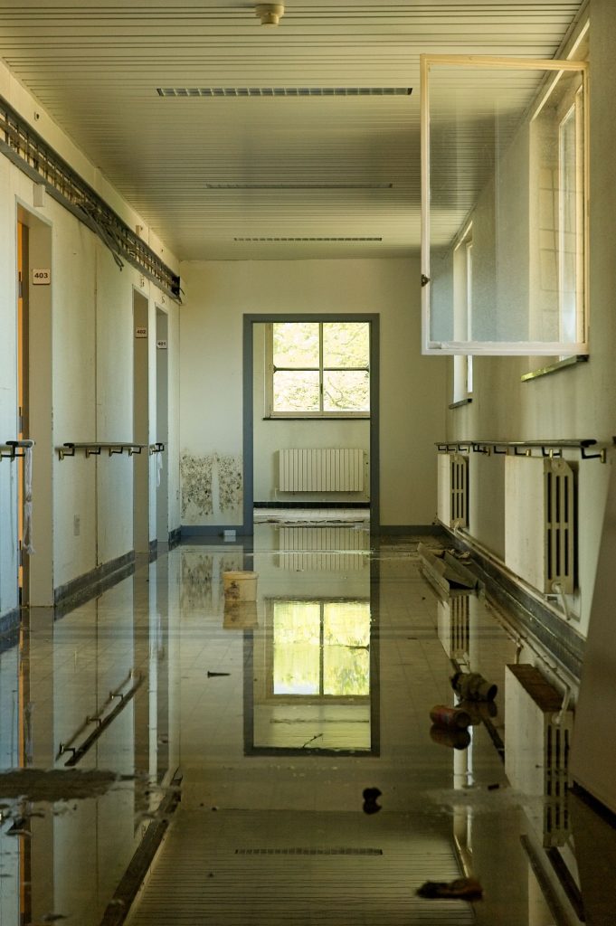 Flooded abandoned room with standing water reflecting a window, representing the hands-full bottleneck on a water damage job site