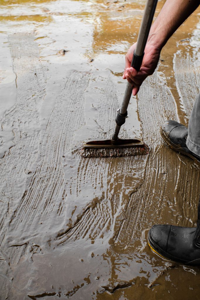 Worker in rubber boots cleaning a muddy floor, showing hands occupied with the physical work of restoration